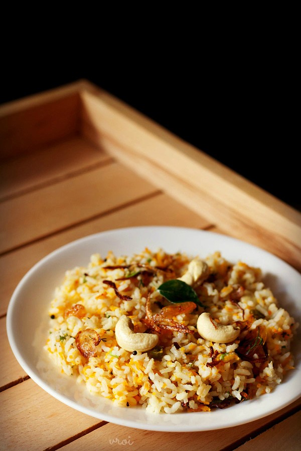 carrot rice garnished with cashews, fried onions and raisins served in a white plate on a light wooden tray