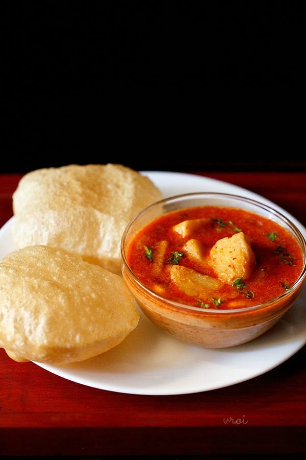 potato kurma served in a glass bowl with pooris kept on left side on a white plate. 