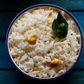 overheat shot of coconut rice topped with two fried curry leaves and cashew bits in a blue rimmed white bowl on a dark blue wooden tray