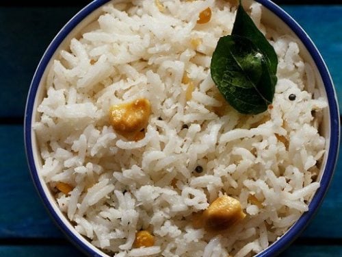 overheat shot of coconut rice topped with two fried curry leaves and cashew bits in a blue rimmed white bowl on a dark blue wooden tray