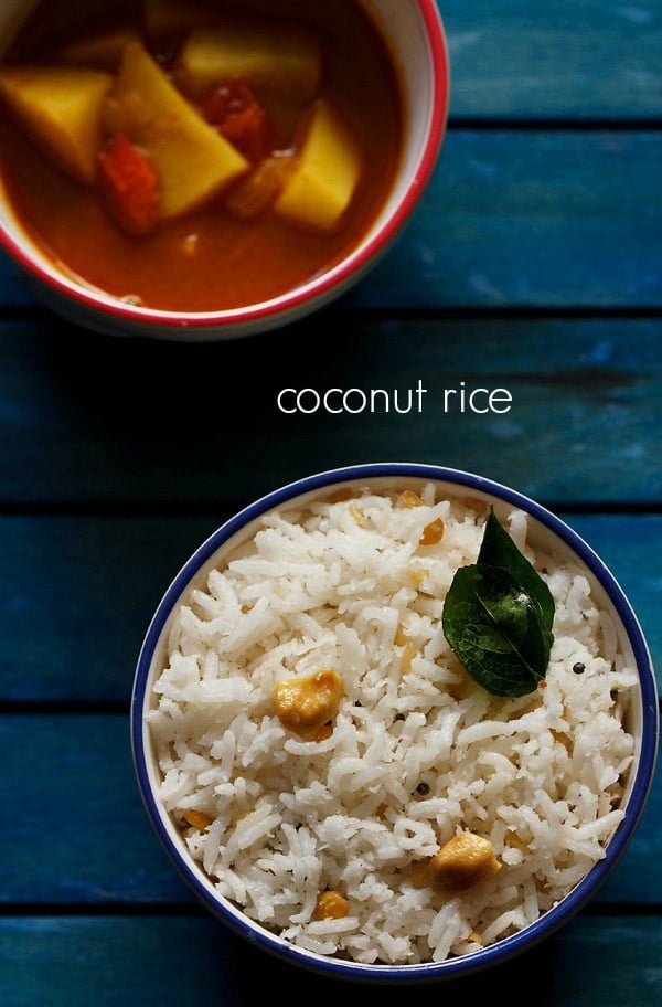 overheat shot of coconut rice topped with two fried curry leaves and cashew bits in a blue rimmed white bowl on a dark blue wooden tray