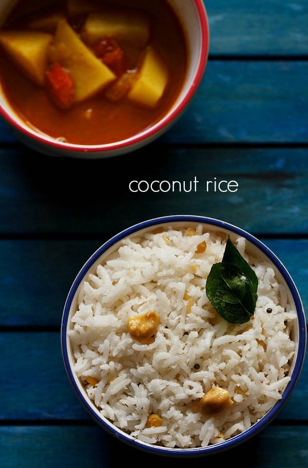 overheat shot of coconut rice topped with two fried curry leaves and cashew bits in a blue rimmed white bowl on a dark blue wooden tray and text layover.