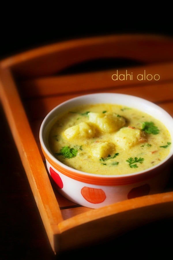 dahi wale aloo garnished with coriander leaves and served in a white with red dots ceramic bowl, kept on a wooden tray and text layover.