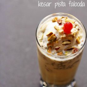 kesar pista falooda garnished with chopped nuts, glazed cherry and served in a tall glass with text layover.
