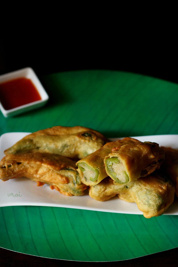 rajasthani mirchi vada served on a white platter with one halved vada on top and a bowl tomato sauce kept on the top left side. 