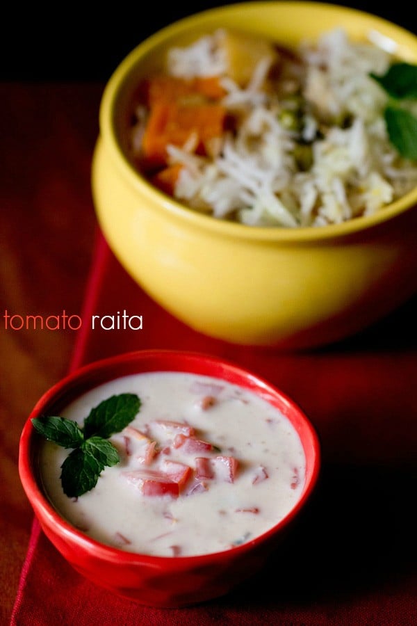tomato raita served in a red bowl with mint leaf sprig for garnish with a yellow bowl of vegetable biryani in the background