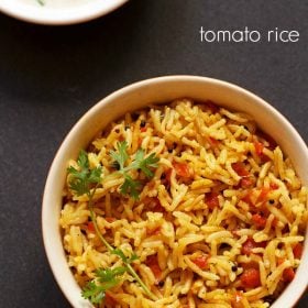 tomato rice with a sprig of cilantro served in an orange and red dotted bowl with a side yogurt raita on a dark black board