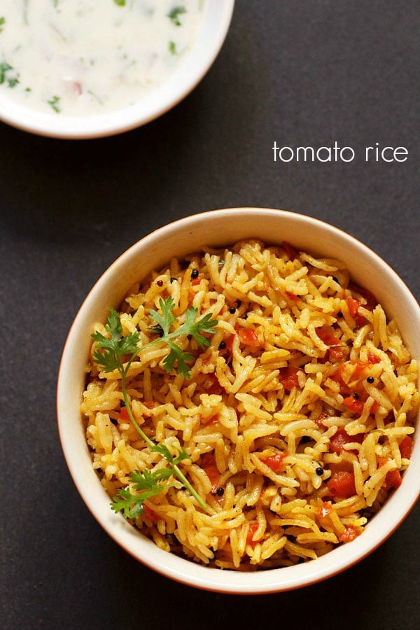 tomato rice with a sprig of cilantro served in an orange and red dotted bowl with a side yogurt raita on a dark black board