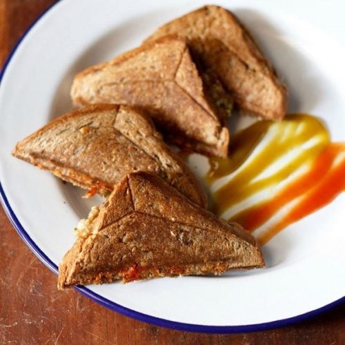 four capsicum sandwich triangles served on a white plate with tomato ketchup and chili sauce.