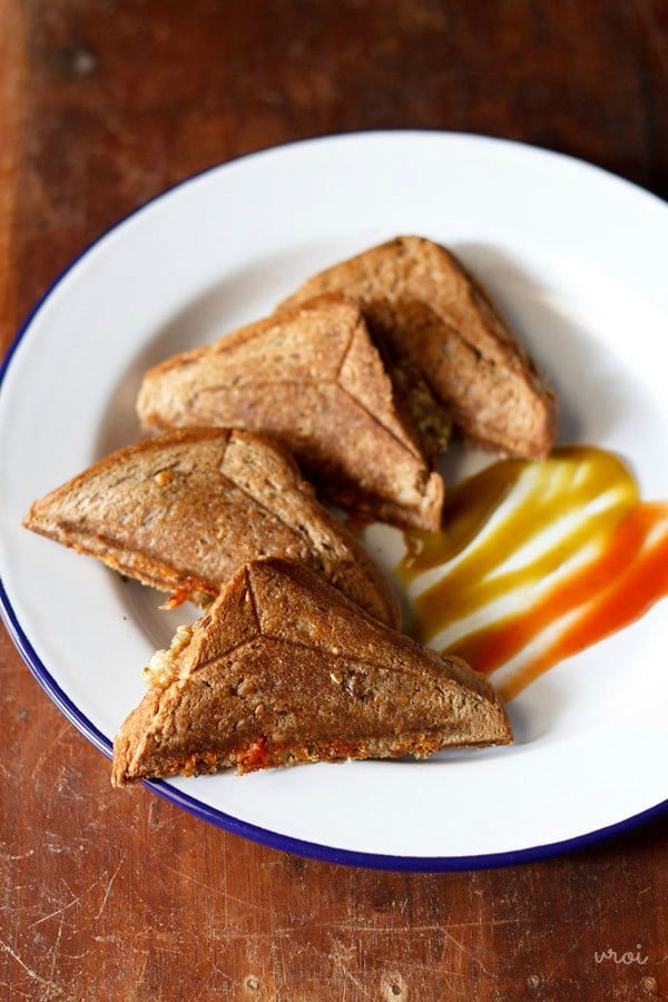 four capsicum sandwich triangles served on a white plate with tomato ketchup and chili sauce. 