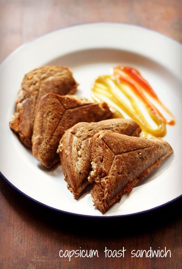 four capsicum sandwich triangles served on a white plate with tomato ketchup and chili sauce.