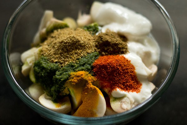 Ground paste, spices, curd, and chopped mushrooms in glass bowl.