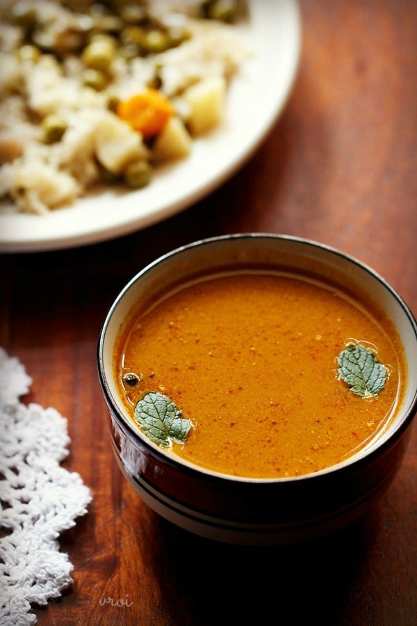 shorba gravy garnished with mint leaves and served in a black rimmed ceramic bowl with a plate of vegetable pulao kept in the background. 