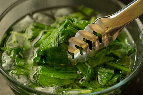 blanched spinach leaves held with a pair of tongs on top of the bowl