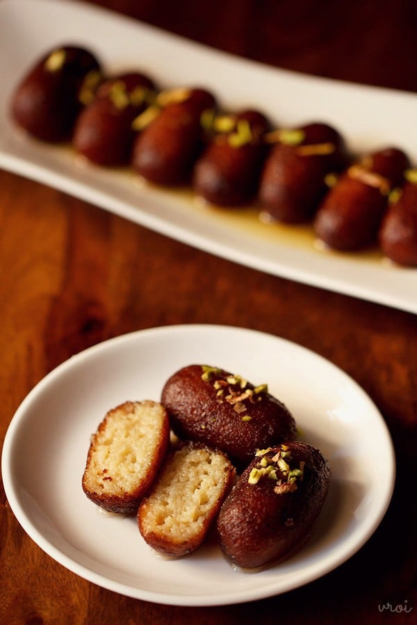 3 kala jamun on a white plate with one cut into half to show the interior texture and the other 2 topped with chopped pistachios.