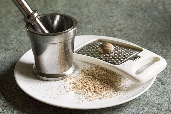 grated nutmeg on a plate with the whole nutmeg and a cheese grater.