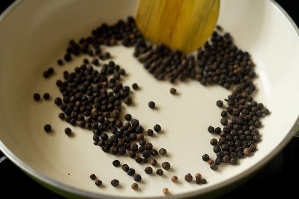 using a wooden spoon to stir whole peppercorns as they are roasting for making sambhar podi