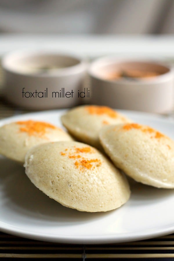 millet idli sprinkled with podi and served on a white plate with a side of bowls of sambar and coconut chutney. 