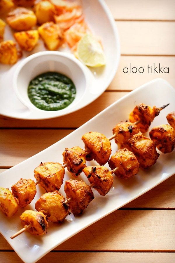 tandoori aloo tikka served on a white rectangular platter with a bowl of green chutney on the top left side and text layovers.