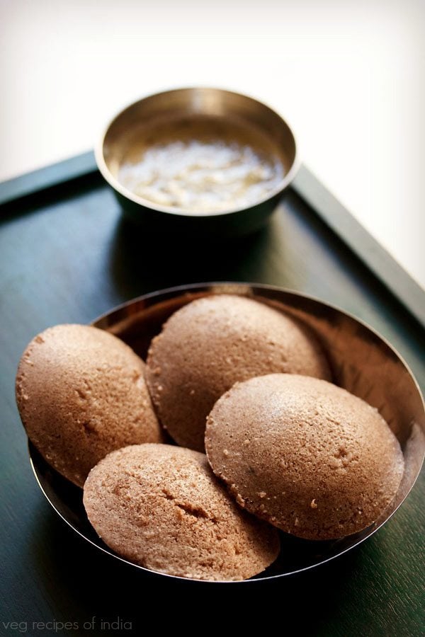 ragi idli served in a steel bowl with a small bowl of coconut chutney kept on the top on a wooden tray with text layovers. 