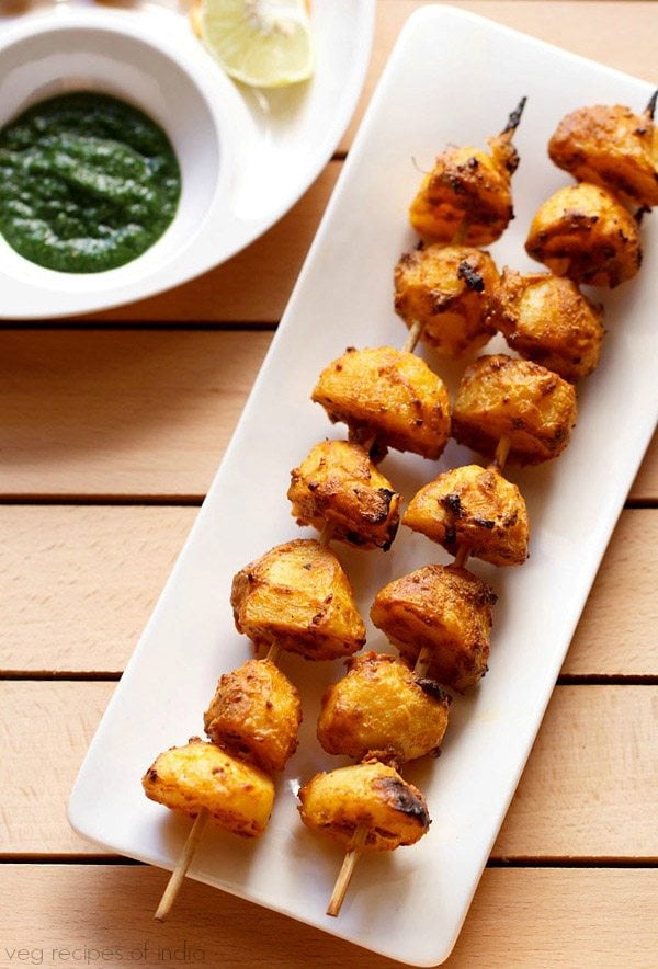 tandoori aloo tikka served on a white rectangular platter with a bowl of green chutney on the top left side and text layover. 