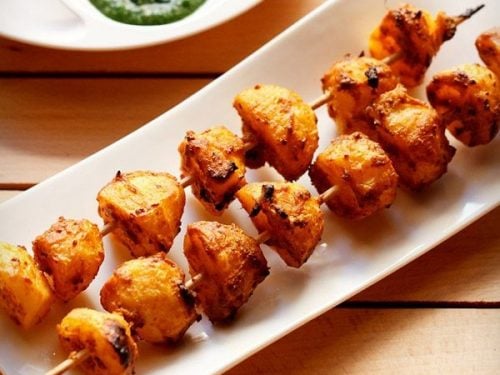 tandoori aloo tikka served on a white rectangular platter with a bowl of green chutney on the top left side and text layovers.