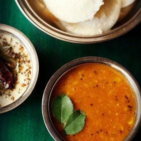 tomato sambar served in a bowl with idlis in a bowl kept on the side, coconut chutney and text layover.