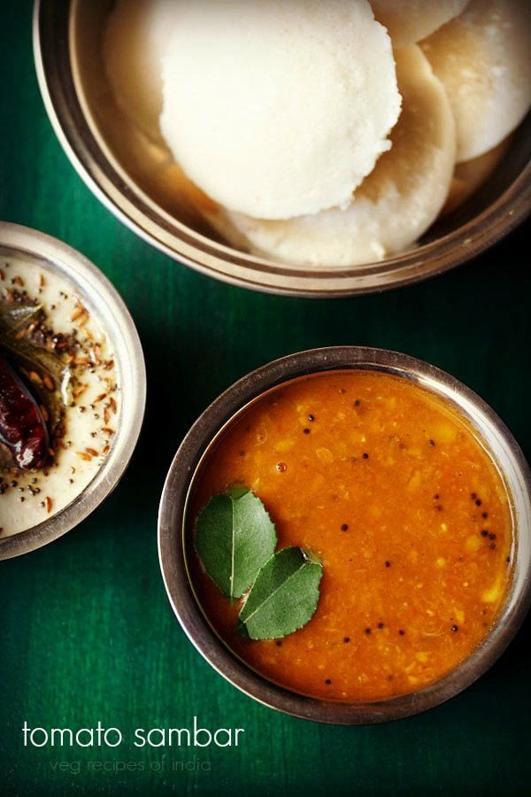 tomato sambar served in a bowl with idlis in a bowl kept on the side, coconut chutney and text layover.