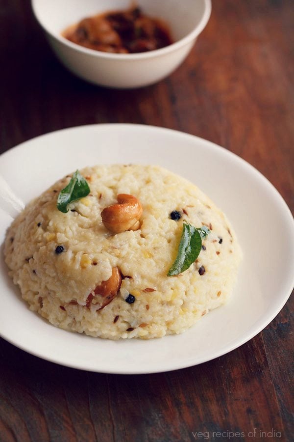 ven pongal garnished with a cashew and curry leaves and served in a white plate with a white bowl of mango pickle by side