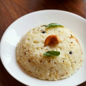 pongal recipe garnished with a cashew and curry leaves and served in a white plate with a white bowl of mango pickle by side