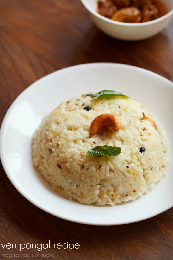 pongal recipe garnished with a cashew and curry leaves and served in a white plate with a white bowl of mango pickle by side