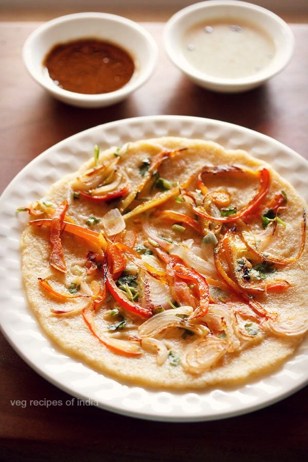 oats uttapam served on a white plate with bowls of chutneys kept in the background. 