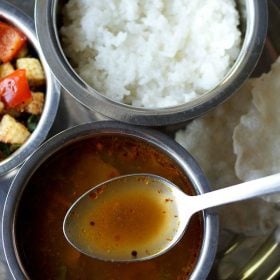 Milagu rasam is served in a steel bowl and a bowl of vegetable stir fry, steamed rice and text layover on a steel spoon on a plate.