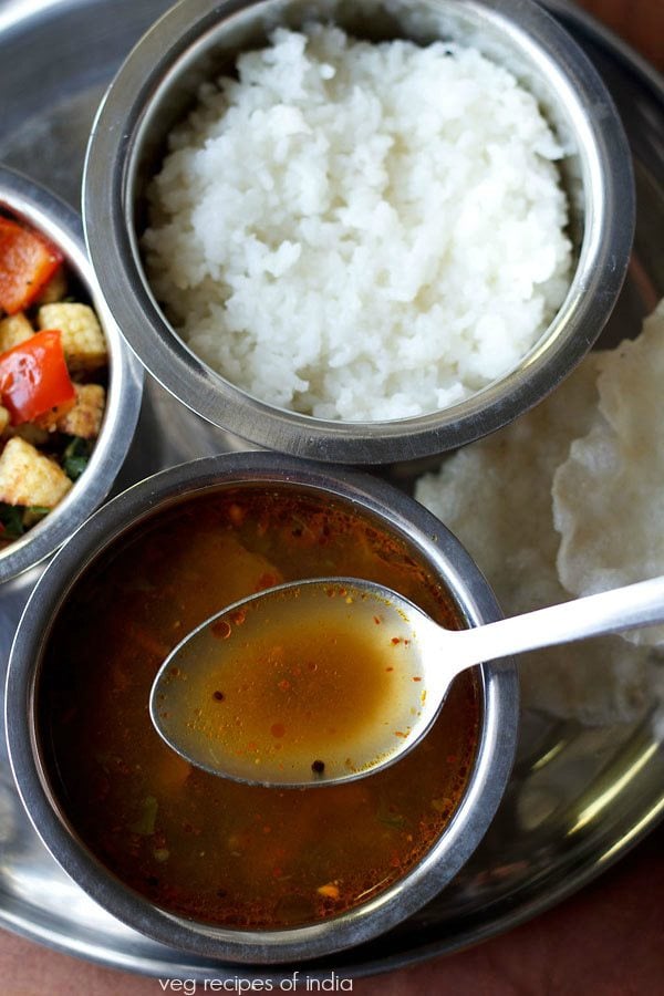 milagu rasam served in a steel bowl with some in a steel spoon on a plate with a bowl of vegetable stir fry, steamed rice and text layover.