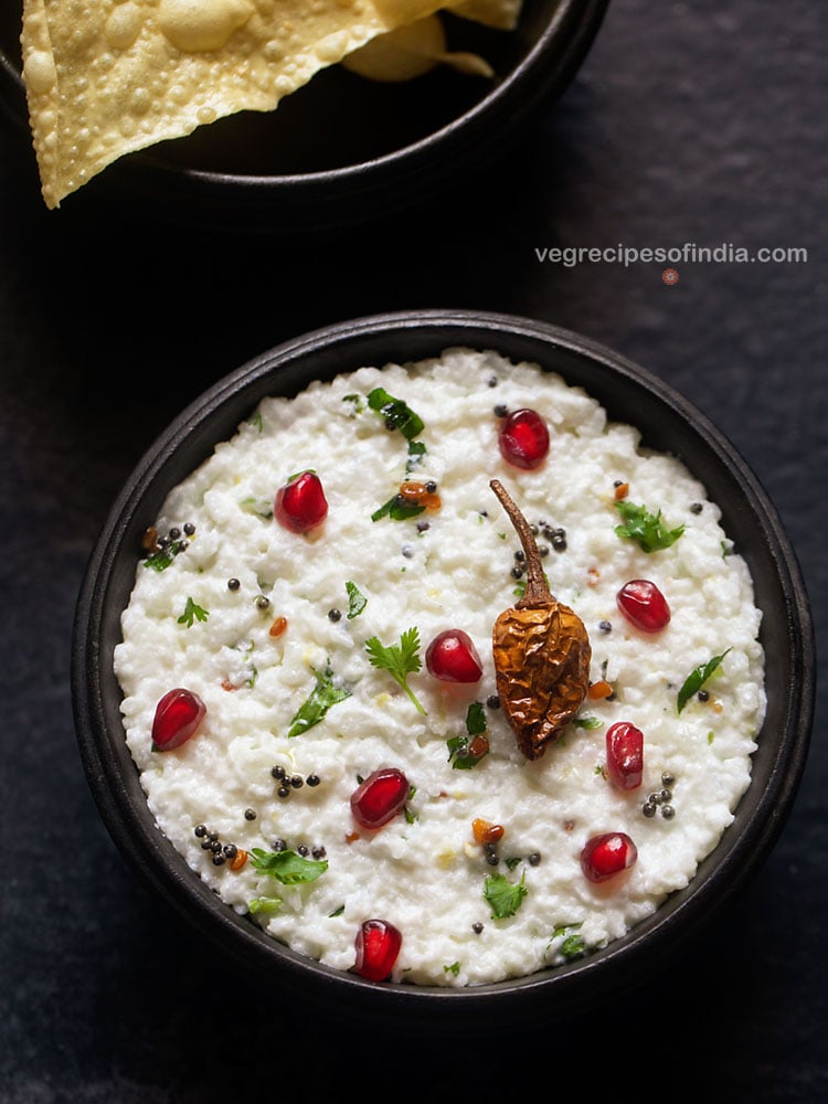 curd rice or daddojanam served in a wooden bowl on a dark grey black board, garnished with chopped coriander leaves, some pomegranate arils and fried sun dried chili. 