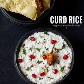 curd rice served in a wooden bowl on a dark grey black board. curd rice is garnished with chopped coriander leaves, some pomegranate arils and fried sun dried chilly. fried papaddum served as as side.
