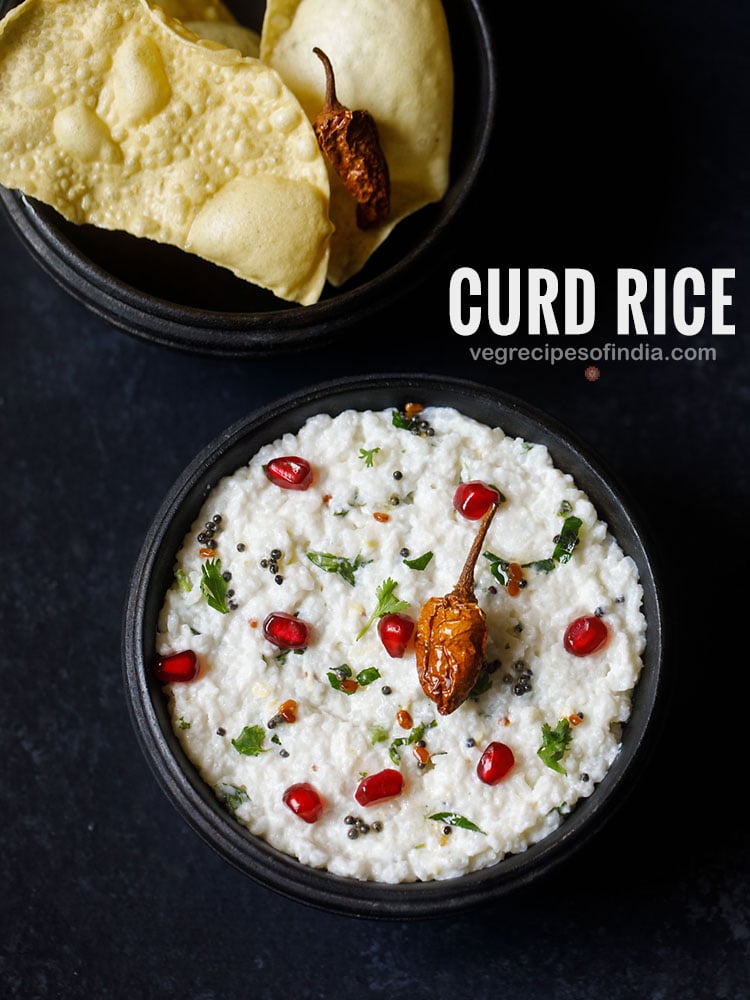 curd rice served in a wooden bowl on a dark grey black board. curd rice is garnished with chopped coriander leaves, some pomegranate arils and fried sun dried chilly. fried papaddum served as as side.