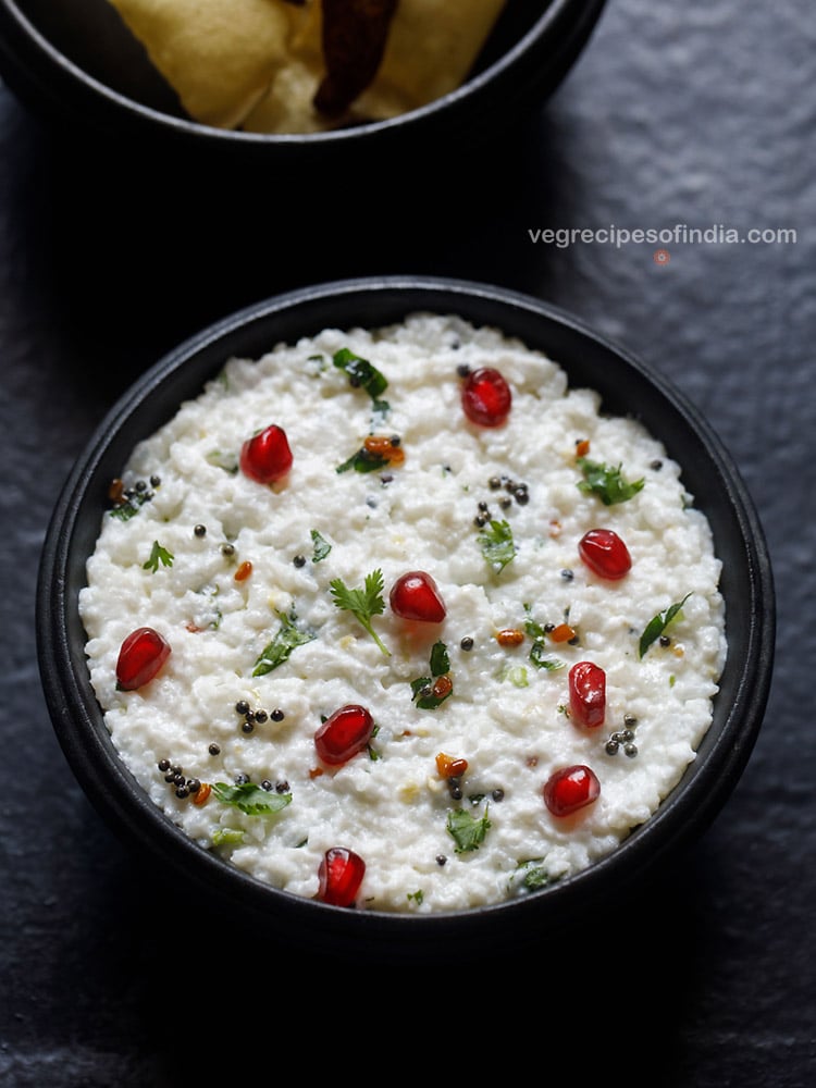 curd rice served in a wooden bowl on a dark grey black board, curd rice is garnished with chopped coriander leaves, some pomegranate arils and fried sun dried chili.