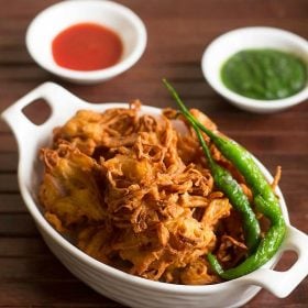 kanda bhaji served in white tray with a side of fried green chilies, green chutney and tomato sauce