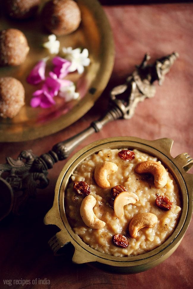 sakkarai pongal served in an antique brass pan with a side of an antique brass spoon and some ladoo and dark pink and white flowers on a brass plate