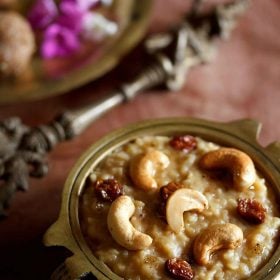 sweet pongal served in an antique brass pan with a side of an antique brass spoon and some ladoo and dark pink and white flowers on a brass plate