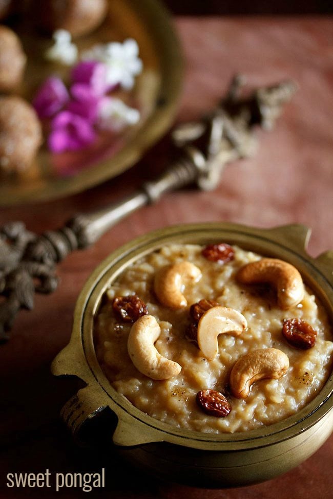 sweet pongal served in an antique brass pan with a side of an antique brass spoon and some ladoo and dark pink and white flowers on a brass plate