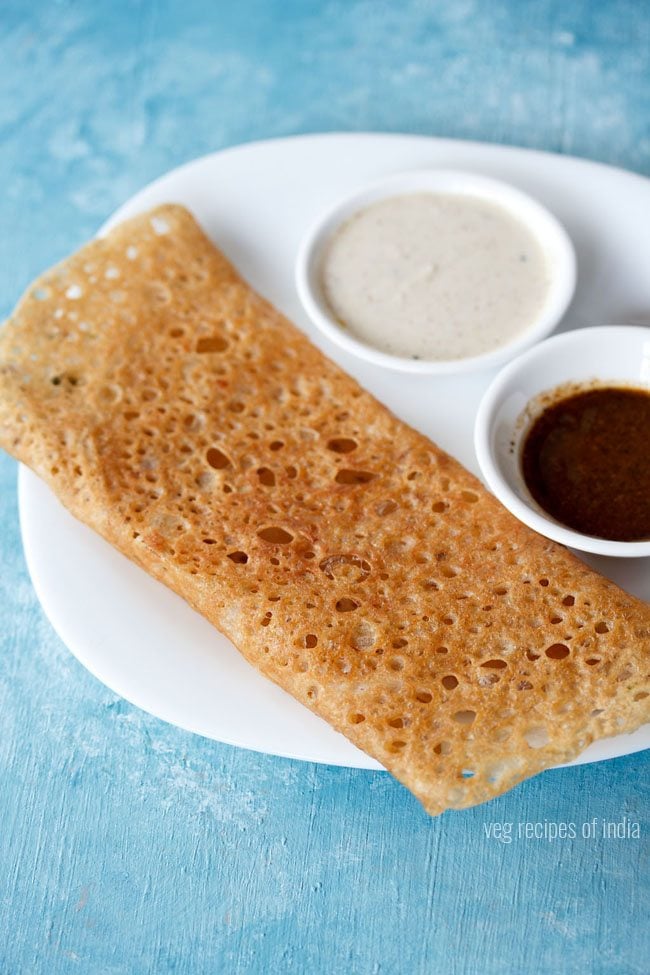 wheat dosa served on a white plate with chutneys in 2 small bowls