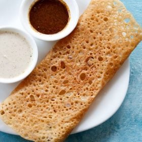 wheat dosa served on a white plate with chutneys in 2 small bowls.