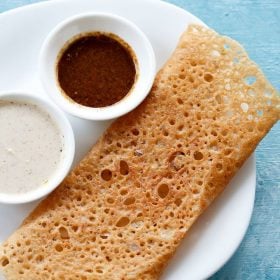 wheat dosa served on a white plate with chutneys in 2 small bowls