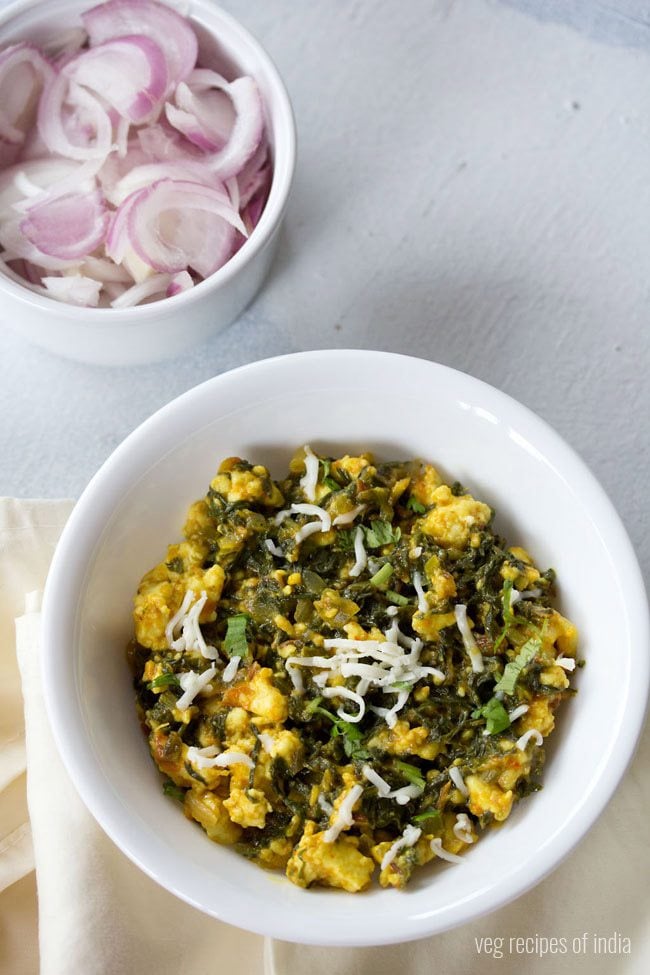 palak paneer bhurji garnished with grated paneer and served in a white bowl along with a side of a bowl of onions. 