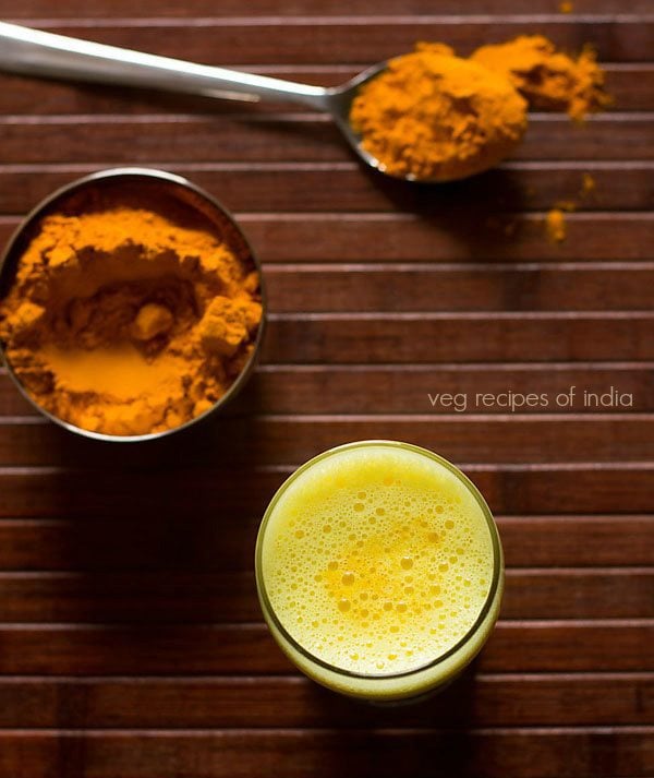 Turmeric milk or golden milk is served in a glass with turmeric in a bowl and spoon on the side.