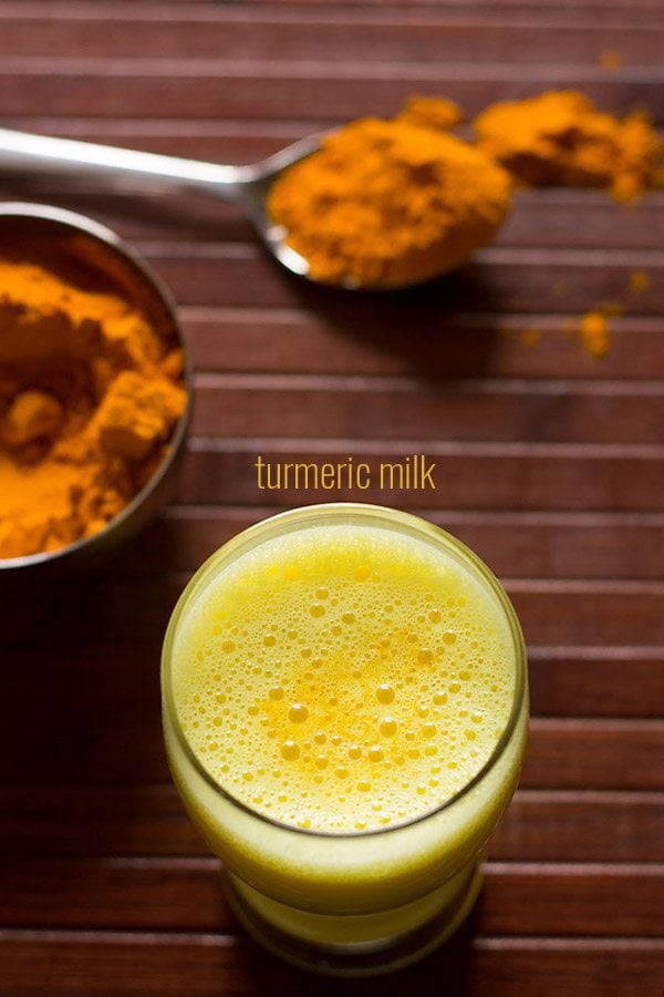 overhead shot of turmeric milk with ground turmeric kept in a bowl and steel spoon on a wooden table.