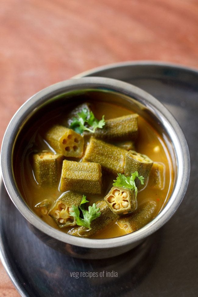 Bendakaya pulusu is garnished with coriander leaves and served in a steel bowl with text in the middle. 