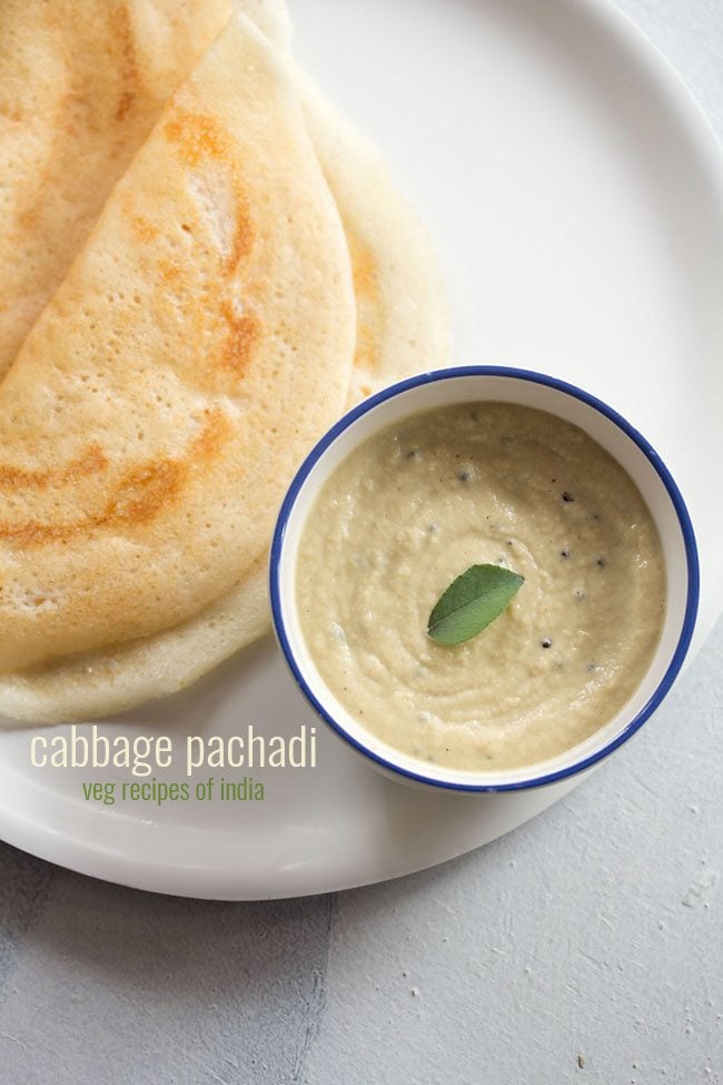 cabbage pachadi with a curry leaf in center in a blue rimmed white bowl with two dosa by the side in a white plate